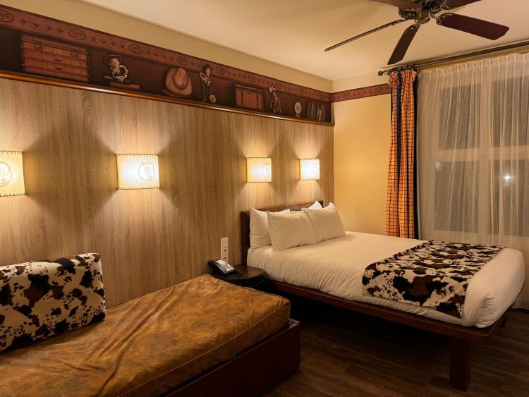 A wider shot of a Hotel Cheyenne room showing a double bed with a cow print bed runner, a pull-out daybed, Western-themed wall decor, and a ceiling fan.