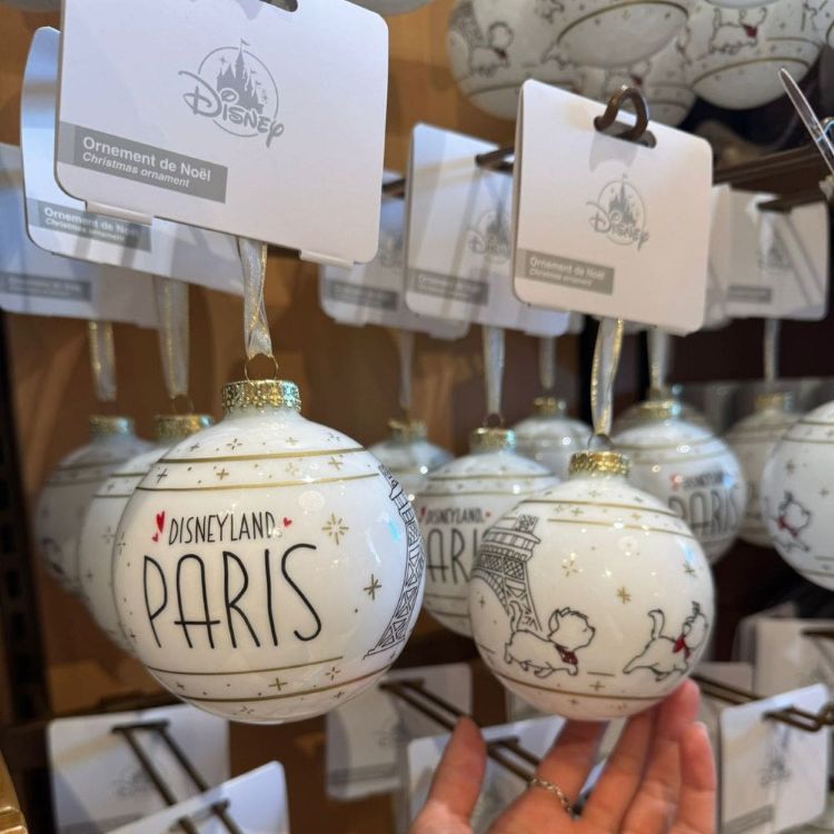A hand holding two white Christmas ornaments, one reading "Disneyland Paris" and the other depicting Parisian landmarks.