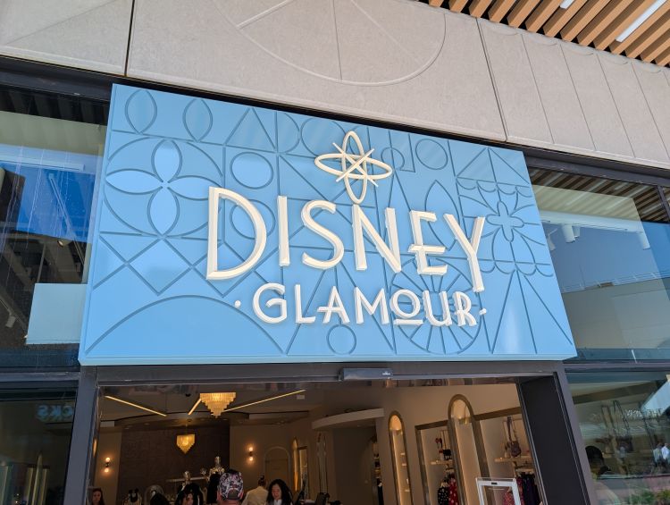 The entrance facade of the Disney Glamour store in Disneyland Paris's Disney Village, featuring a large light blue sign with white "DISNEY GLAMOUR" lettering.