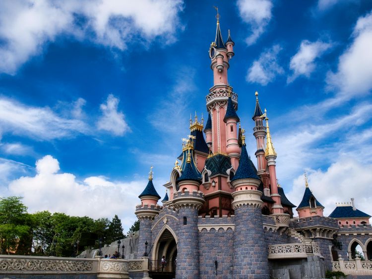 The Sleeping Beauty Castle at Disneyland Paris, a pink and blue fairy-tale castle with spires, under a bright blue sky with scattered clouds.