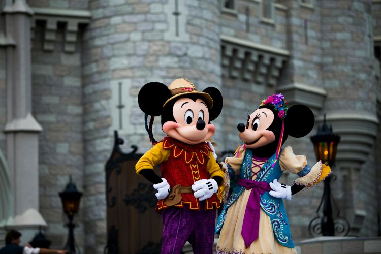 Mickey Mouse and Minnie Mouse in elaborate, colourful costumes standing in front of a stone castle facade. Mickey wears a red and gold tunic with a feathered cap, and Minnie wears a light blue and purple dress with a floral headpiece.