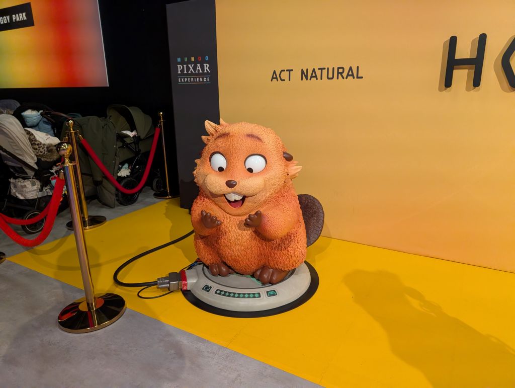 A life-size statue of Mabel the beaver from the movie Hoppers, standing on a grey base in front of a yellow "ACT NATURAL" wall at the Pixar Mundo entrance.