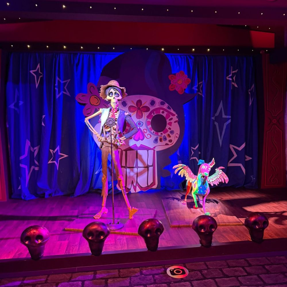 A vibrant stage set from the movie Coco featuring a life-size figure of Hector with his guitar and the spirit guide Dante against a backdrop of a large decorative skull and blue curtains with stars.