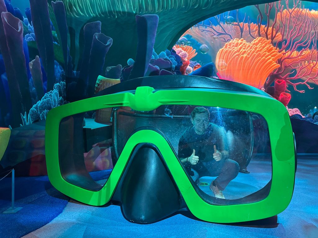A man posing with two thumbs up behind a giant green diver’s mask in a Finding Nemo themed room with colorful coral reef backdrops.