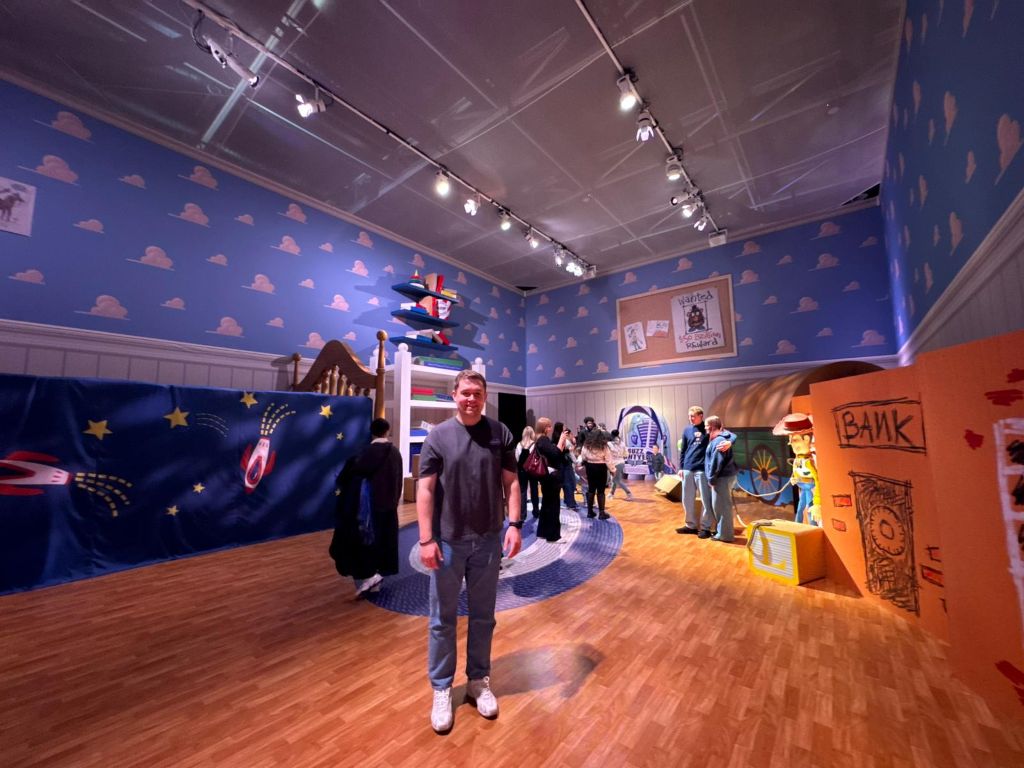 A wide-angle view of a man standing in a life-size recreation of Andy’s bedroom from Toy Story, featuring oversized blue cloud wallpaper and giant furniture to make guests feel toy-sized.