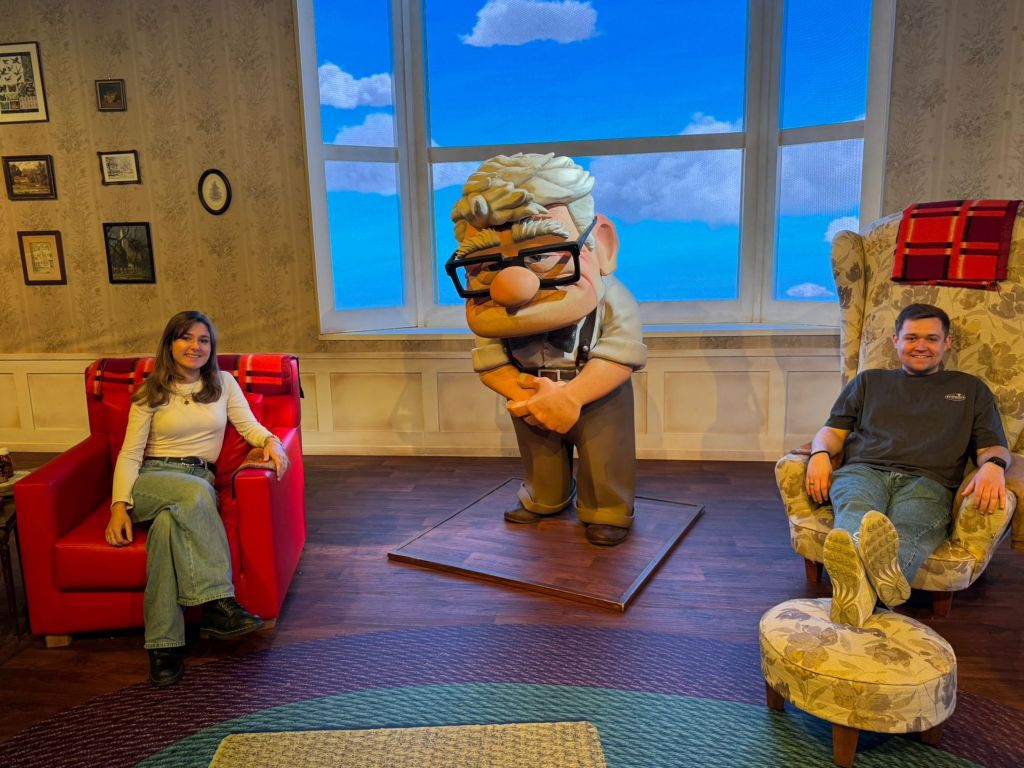 Two guests sitting in Carl and Ellie’s famous armchairs next to a large figure of Carl Fredricksen in a room that recreates the living room from Pixar's Up.
