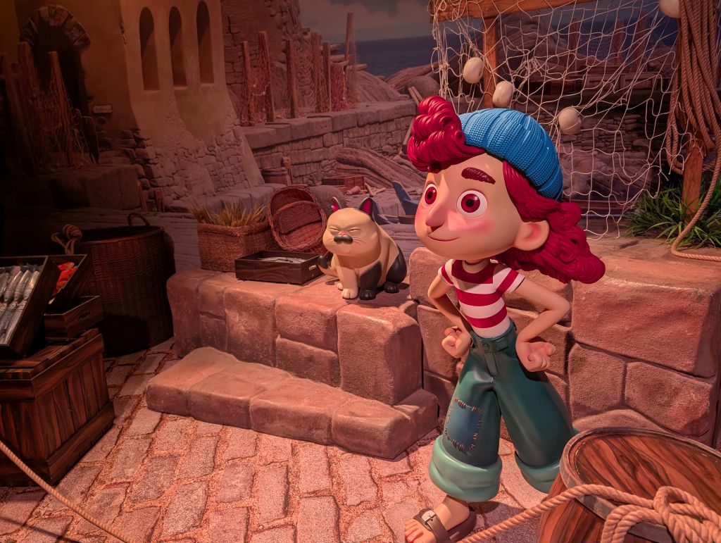 A life-size figure of Giulia Marcovaldo standing in a recreated Italian harbor scene from the movie Luca, featuring stone steps, fishing nets, and the cat Machiavelli sitting on a ledge.