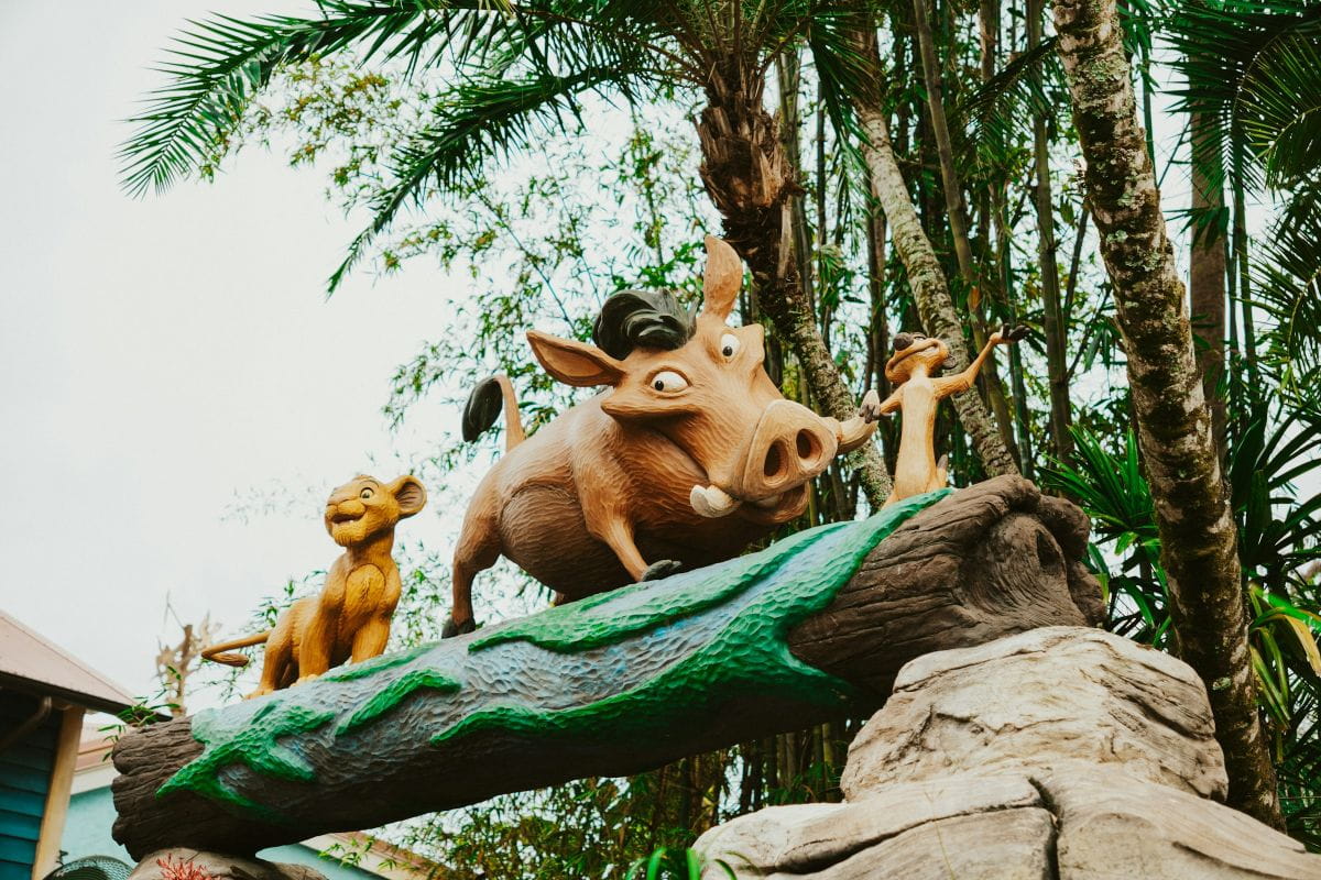 A vibrant outdoor statue of Simba, Pumbaa, and Timon from Disney's The Lion King walking across a log, surrounded by lush tropical palm trees.