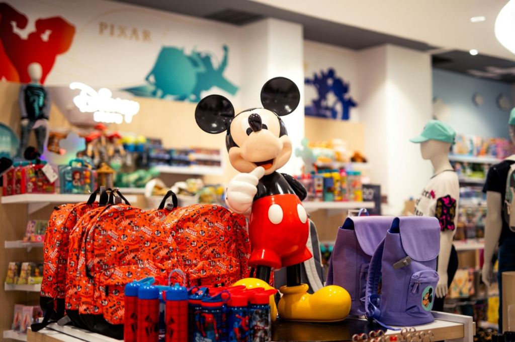 A vibrant Disney Store display featuring a large Mickey Mouse statue surrounded by backpacks, water bottles, and classic Pixar character posters in the background.