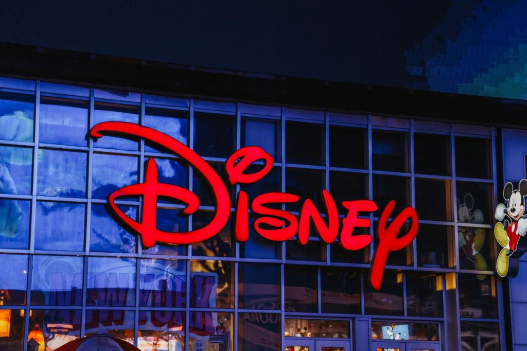 A close-up of the bright red Disney logo on a glass storefront, reflecting the surrounding city lights with a classic Mickey Mouse figure visible on the right.