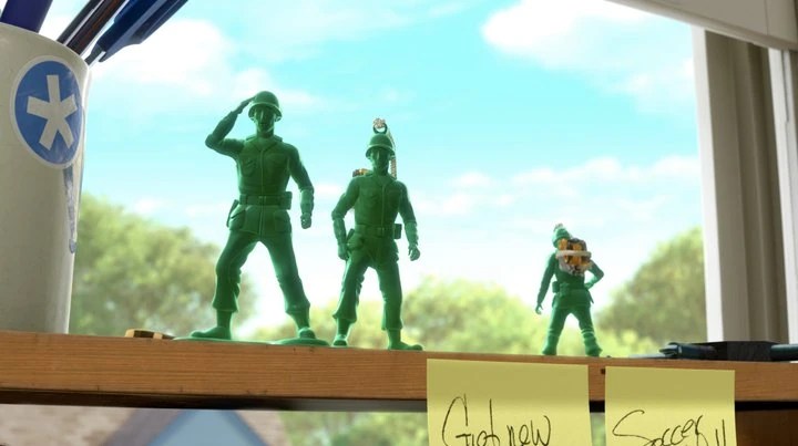 Three green plastic army men toys standing on a wooden shelf near a window, with one soldier saluting against a background of green trees and blue sky.
